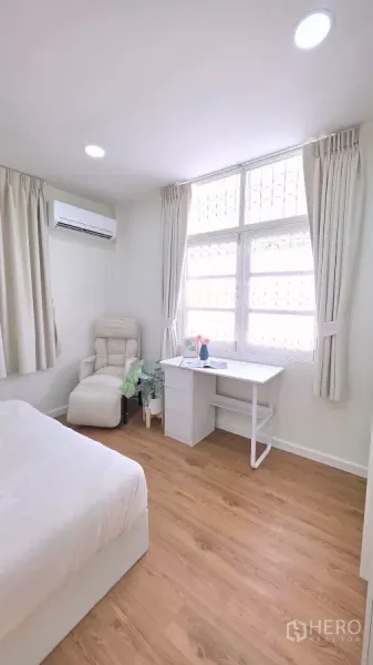 Detached House for rent in Huai Khwang, Bangkok - Sunlit bedroom with desk, armchair and large window with security grilles.