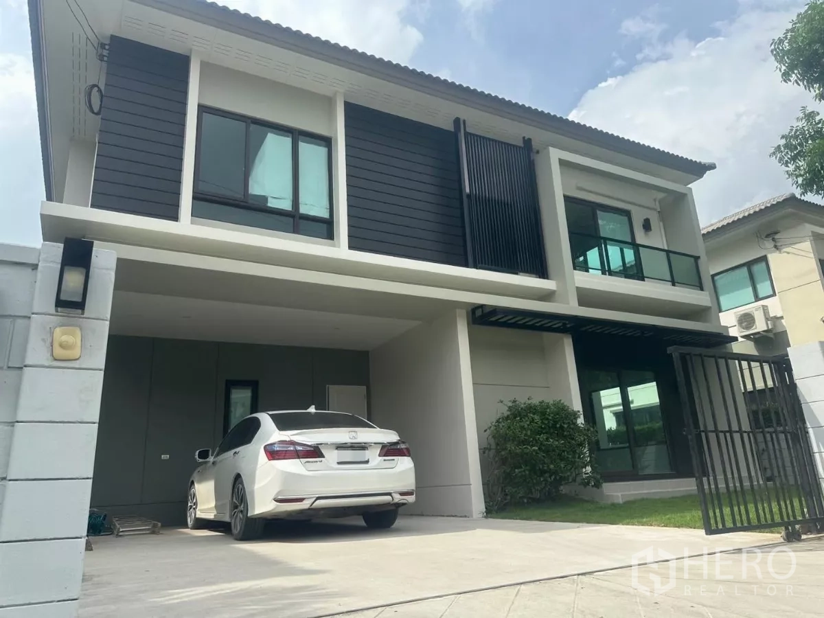 独立式住宅 出售在Samut Prakan - 两层独栋别墅外观，带双车位有顶车棚、大门与前草坪。