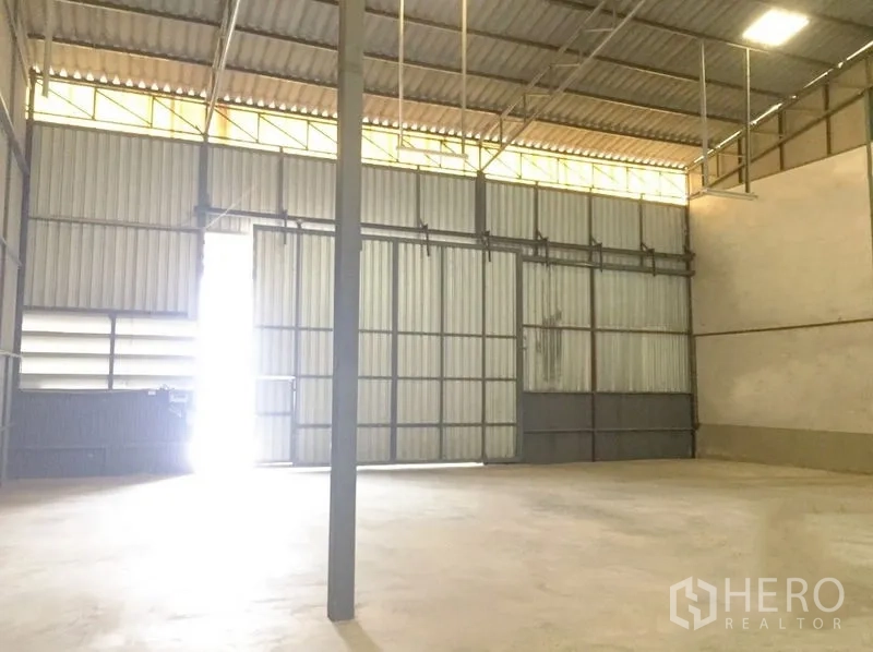 Warehouse for rent in Prawet, Bangkok - High-ceiling warehouse showing steel trusses, skylight panels, and a wide entry door with natural light.