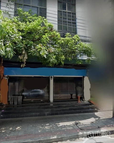 店屋 出租在Watthana, Bangkok - 街边商铺外立面，宽大玻璃门面、台阶入口与浓密树荫，位于瓦他那区。