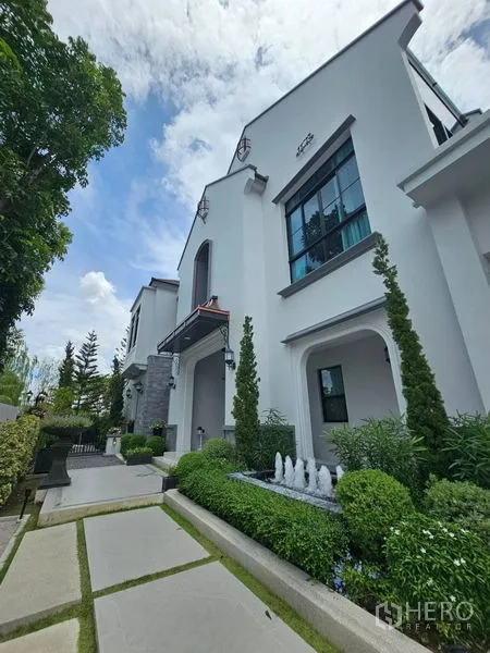 Detached House for rent or sale in Saphan Sung, Bangkok - Elegant white detached house exterior with manicured garden, paved path and a small fountain beneath a blue sky.