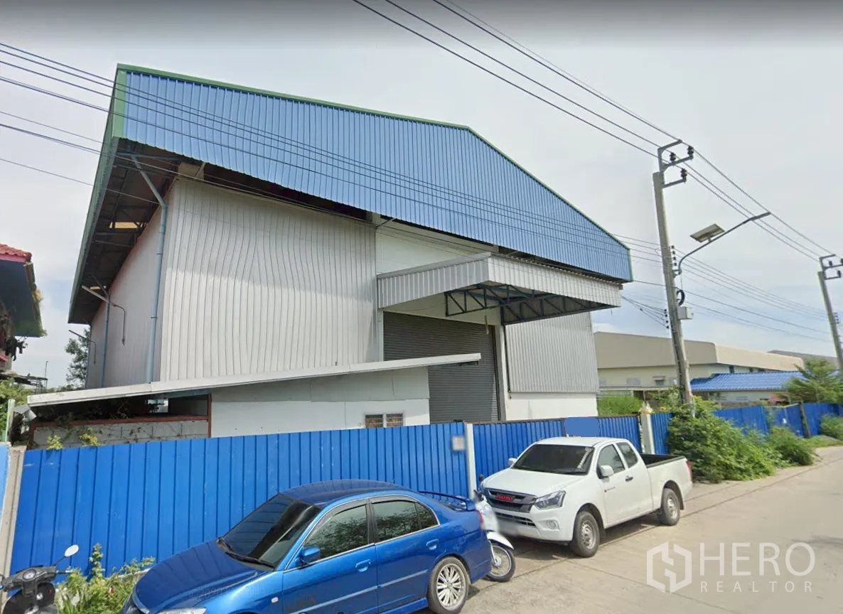 คลังสินค้า ให้เช่าในPak Kret, Nonthaburi - มุมมองด้านหน้าโกดังโครงเหล็ก หลังคาสีฟ้า มีประตูม้วนกว้าง พร้อมกันสาดลานขนถ่ายและรั้วรอบ ปากเกร็ด นนทบุรี
