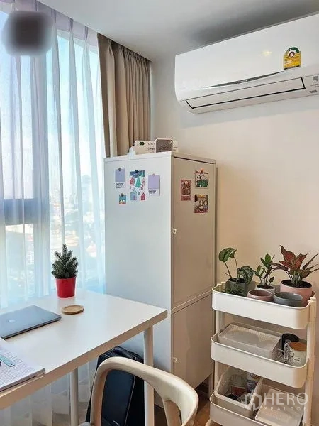 Condominium for rent in Huai Khwang, Bangkok - Compact work-from-home corner by the window with desk, storage cart, plants and air conditioner.