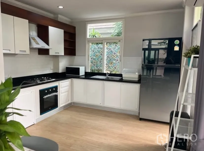 Detached House for rent or sale in Lat Phrao, Bangkok - Modern L-shaped kitchen with white cabinets, black countertop, oven, hob, hood and fridge.