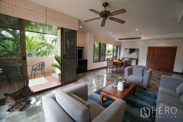 Apartment for rent in Watthana, Bangkok - Open-plan living room with gray sofas, ceiling fan, marble floors, and sliding doors to a leafy balcony.