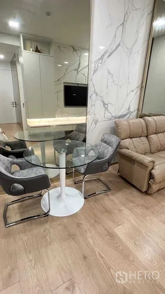Condominium for rent in Watthana, Bangkok - Open-plan dining corner with round glass table and sofa beside a marble accent wall.