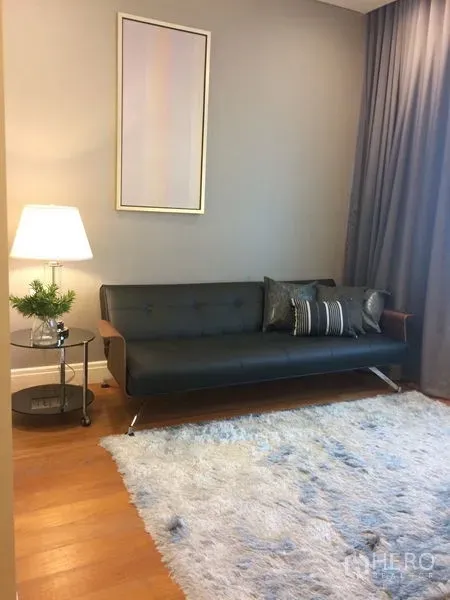 Condominium for rent in Khlong Toei, Bangkok - Compact sitting room with a black sofa, side table and lamp, and a soft white rug.