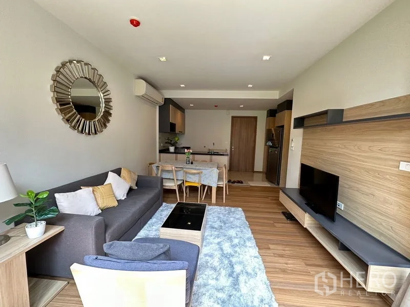 Condominium for rent in Watthana, Bangkok - Spacious living area with sofa, TV wall unit, soft rug, and balcony windows beside the open kitchen.