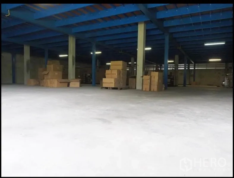 Warehouse for rent in Nakhon Pathom - Open-plan warehouse interior with blue steel beams, concrete floor, and pallets of boxes.