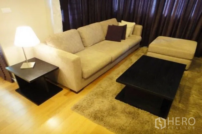 คอนโด ให้เช่าในSathon, Bangkok - ห้องนั่งเล่นอบอุ่น โซฟาสีเบจ ออตโตแมน พรมฟู โต๊ะกาแฟสีเข้ม และผ้าม่านเต็มบาน