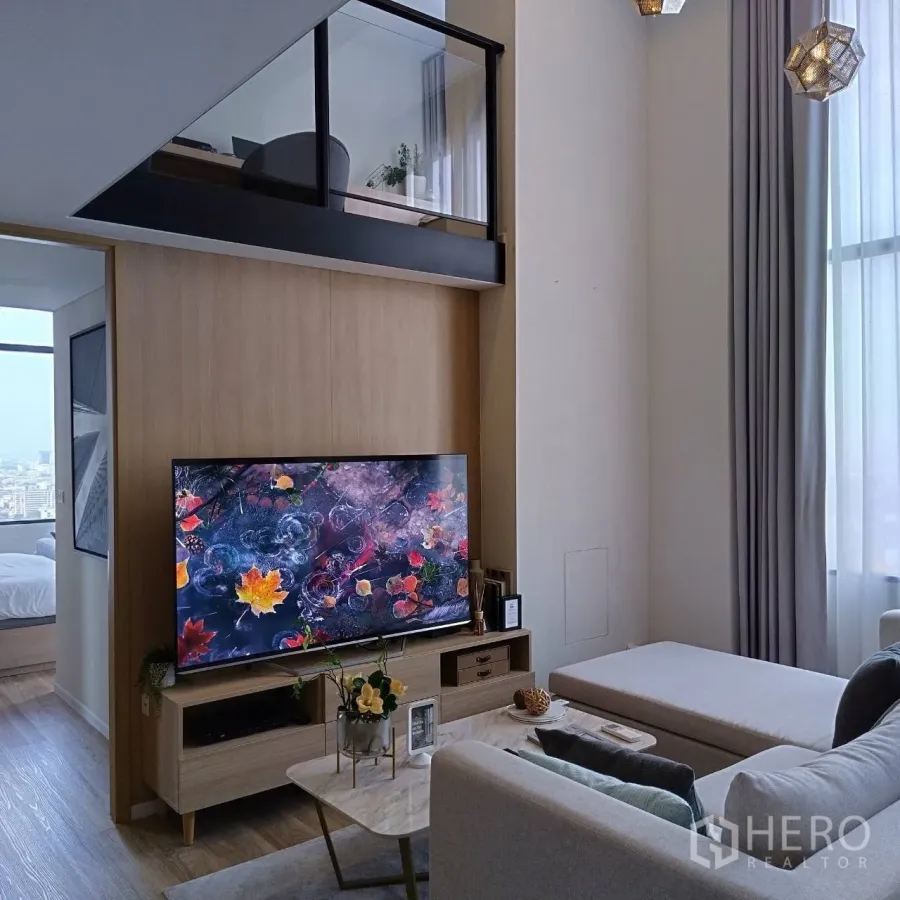 Condominium for rent or sale in Khlong Toei, Bangkok - Double-height lounge with large TV, neutral sofa, and tall windows beneath the mezzanine.