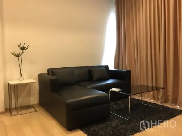 Condominium for rent in Khlong Toei, Bangkok - Contemporary lounge with black L-shaped sofa, glass coffee table, beige curtains, and rug on wood floor.