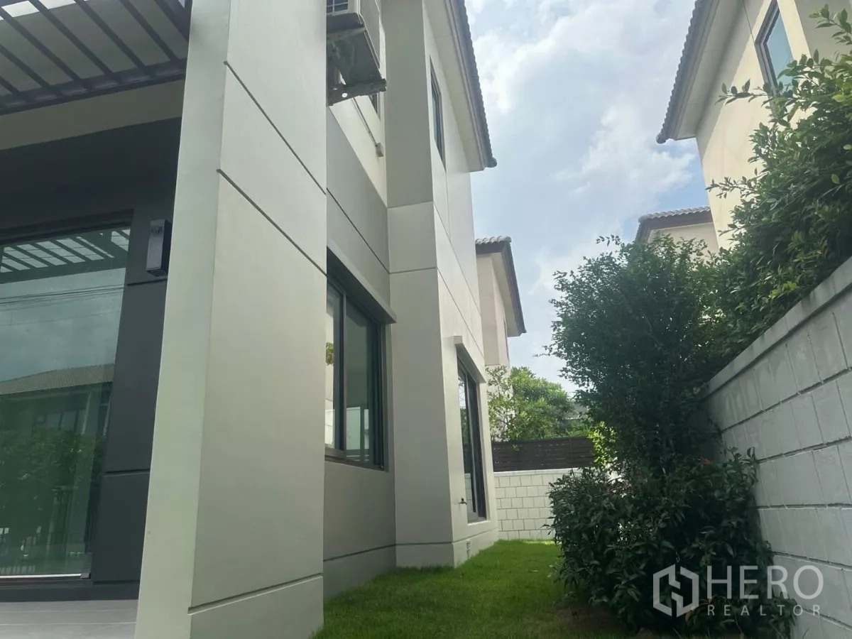 独立式住宅 出售在Samut Prakan - 房屋侧立面，粗柱与大窗，沿墙有草坪与灌木。