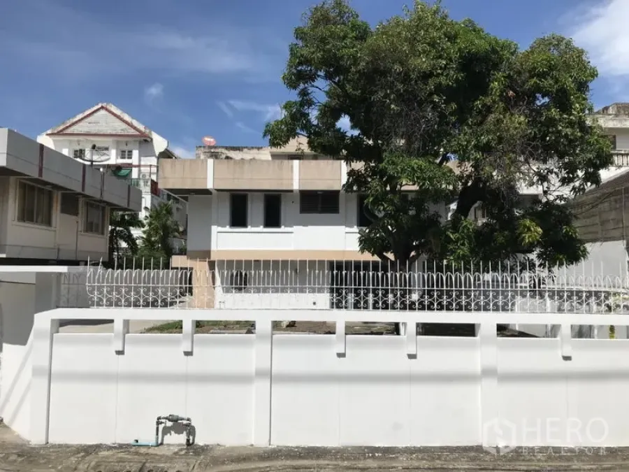 独立式住宅 出租或出售在Khlong Toei, Bangkok - 两层白色独栋住宅，周围有围墙与车道入口，庭院大树遮荫，位于空堤区，天空晴朗。