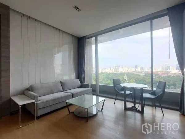 公寓 出租在Bang Rak, Bangkok - 现代客厅，灰色沙发、玻璃茶几和圆形餐桌，旁有全景落地窗与城市景观。