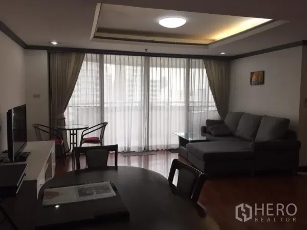 คอนโด ให้เช่าในWatthana, Bangkok - ห้องนั่งเล่นสว่าง โซฟา L‑shape ทีวี มุมทานข้าว และกระจกสูงจรดเพดานเปิดสู่ระเบียงวิวเมือง
