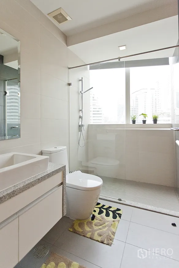 Condominium for rent in Khlong Toei, Bangkok - Contemporary bathroom with glass-enclosed shower, window light, and vanity.