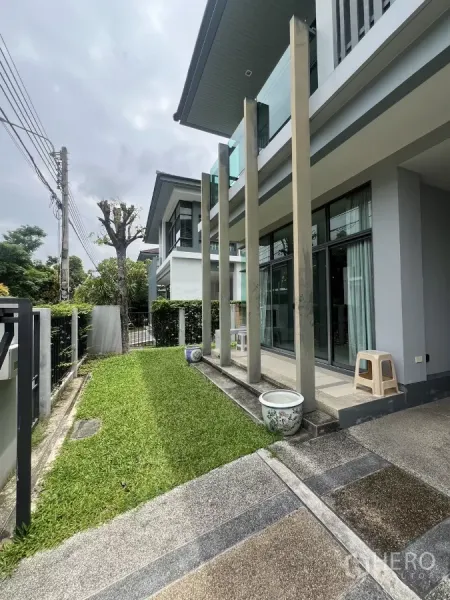 Detached House for rent in Bang Kapi, Bangkok - Side view of the house showing a small lawn, tall columns, and floor-to-ceiling windows.