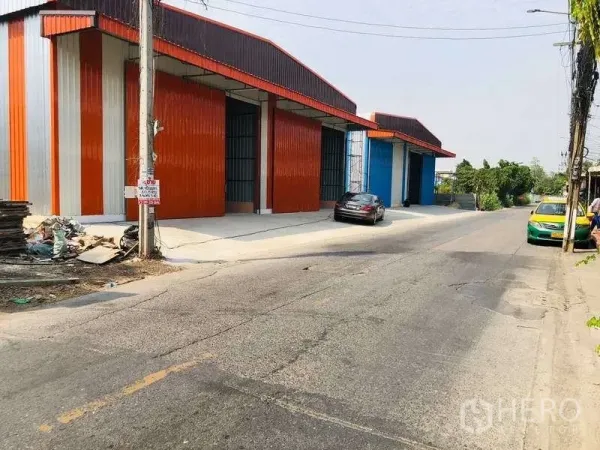 Warehouse for rent in Bueng Kum, Bangkok - Street-side view of colorful steel-clad warehouse units with wide loading aprons and parked cars in Bueng Kum.