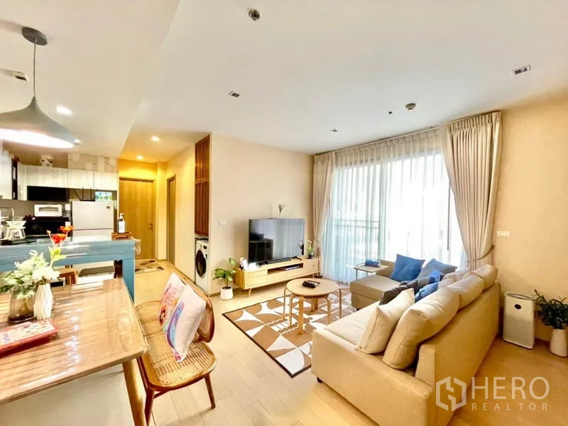 Condominium for rent or sale in Watthana, Bangkok - Bright Thonglor condo living room with L‑shaped sofa, TV, balcony doors and open kitchen bar.