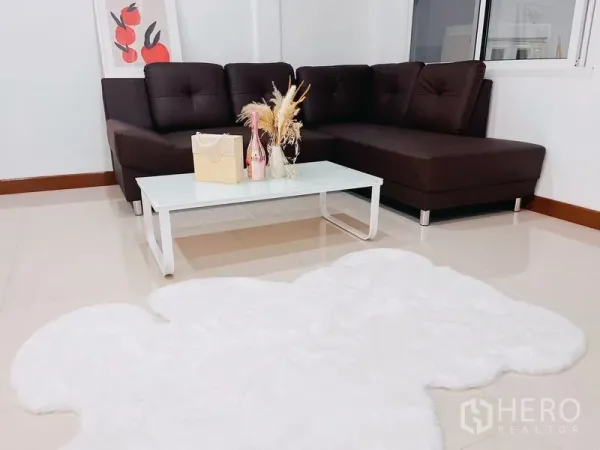 Townhouse for rent in Huai Khwang, Bangkok - Cozy living room with a brown L-shaped sofa, glass coffee table and fluffy white rug.