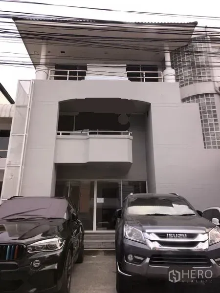 Home Office for rent in Huai Khwang, Bangkok - Three-storey home office exterior with a small front balcony, glass-block corner wall, and parking space with cars in front.