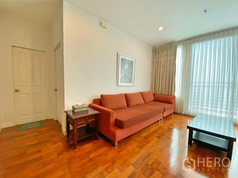 Condominium for rent in Khlong Toei, Bangkok - Bright living room with orange sofa, coffee table and floor-to-ceiling sheer curtains.