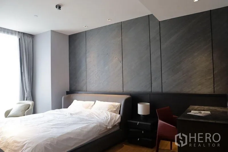 Condominium for rent in Khlong Toei, Bangkok - Modern bedroom with dark textured wall panels, upholstered bed, bedside table and desk, lit by a tall window with curtains.