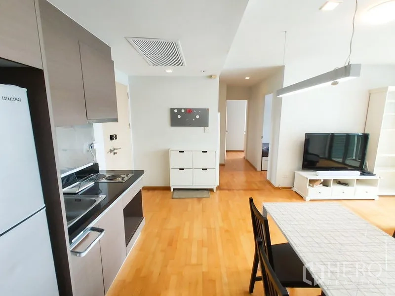 Condominium for rent in Khlong Toei, Bangkok - Compact kitchen with upper cabinets, sink, cooktop and fridge beside dining table.