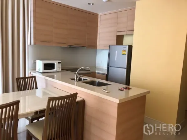 Condominium for rent or sale in Khlong Toei, Bangkok - Open-plan kitchen with light wood cabinets, breakfast bar with sink, and dining set.