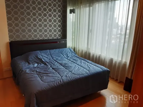 Condominium for rent in Khlong Toei, Bangkok - Bright bedroom with a large window, city views, patterned feature wall, and a queen bed with blue bedding.