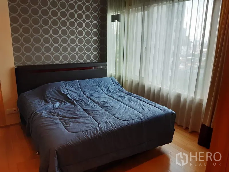 Condominium for rent in Khlong Toei, Bangkok - Bright bedroom with a large window, city views, patterned feature wall, and a queen bed with blue bedding.