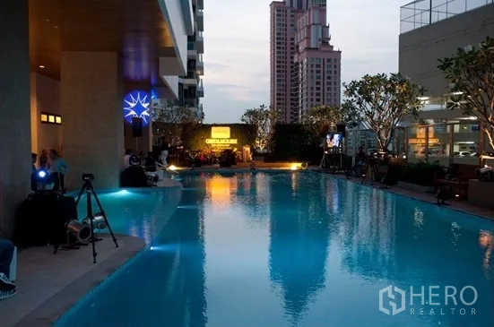 Condominium for rent or sale in Khlong Toei, Bangkok - Condominium outdoor swimming pool at dusk with city backdrop and seating area.