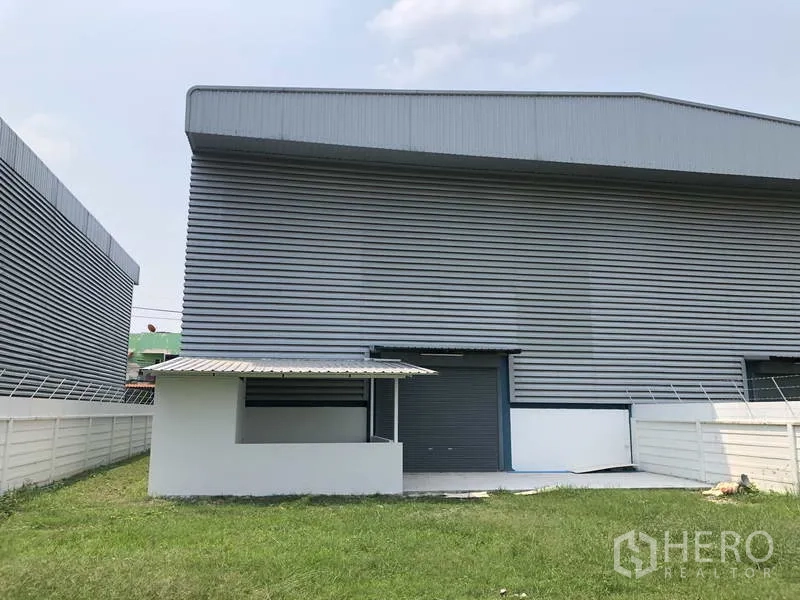 Factory for sale in Sai Noi, Nonthaburi - Rear elevation of the factory with a roller shutter door and small covered service area next to a lawn.