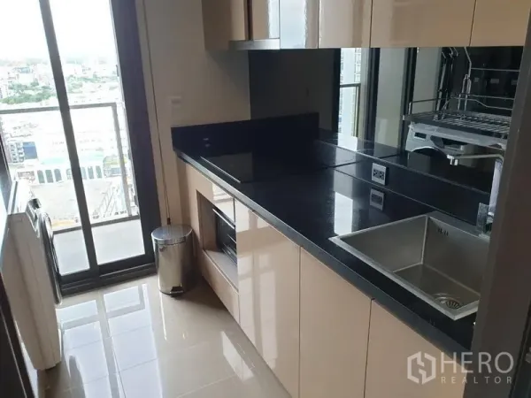 公寓 出租在Watthana, Bangkok - 一字型厨房，亮面橱柜、黑色台面与水槽，阳台门外可见城市景观。