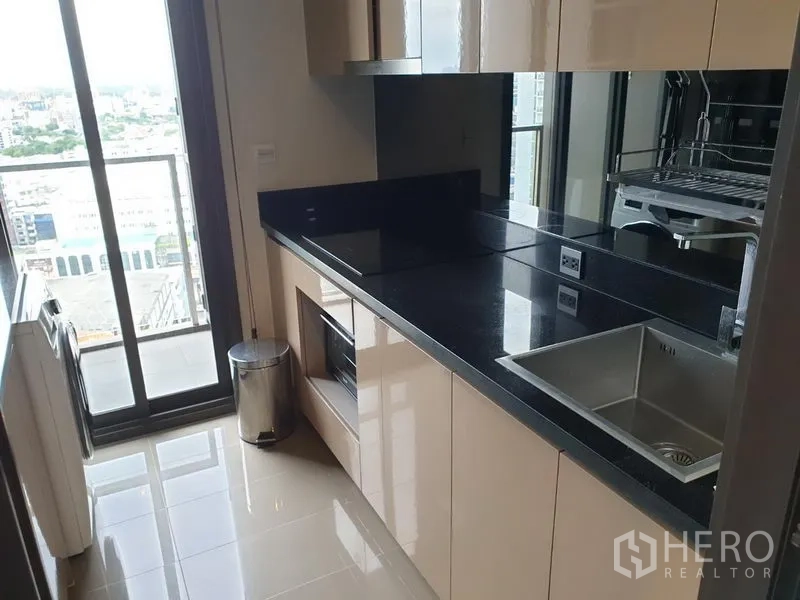 公寓 出租在Watthana, Bangkok - 一字型厨房，亮面橱柜、黑色台面与水槽，阳台门外可见城市景观。