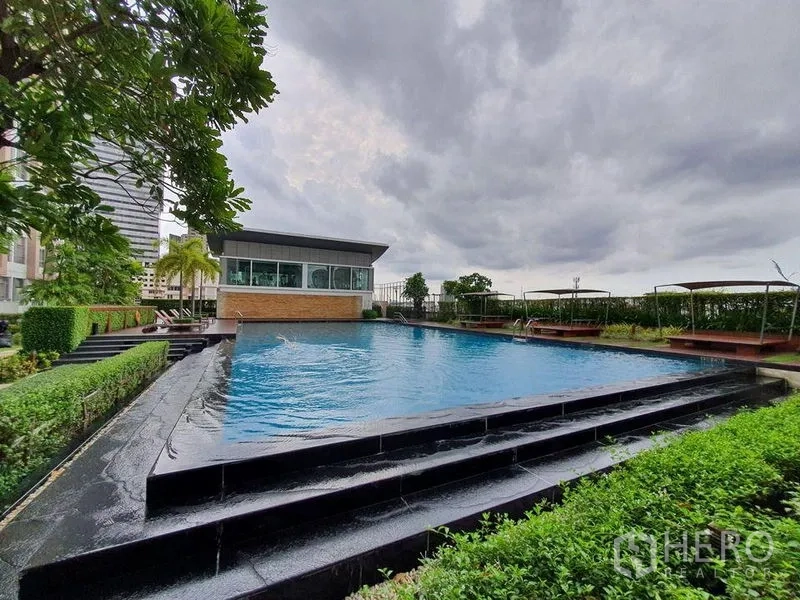 Condominium for sale - Outdoor infinity-edge swimming pool with landscaped gardens and sun decks under a cloudy sky.