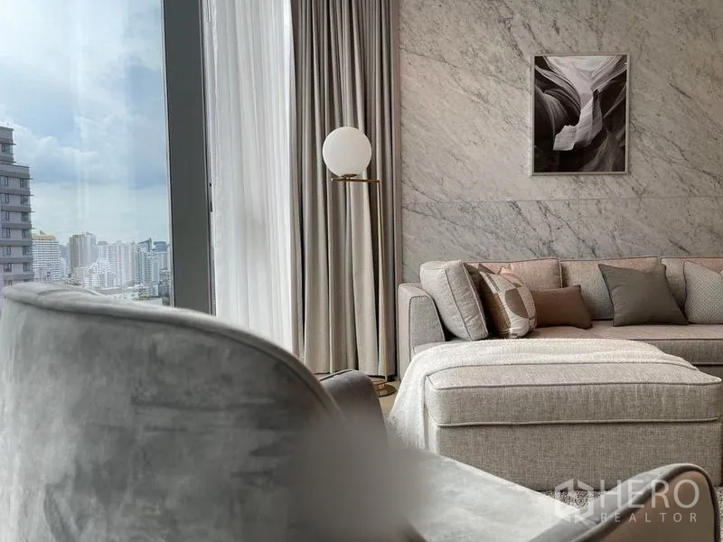 Condominium for rent in Watthana, Bangkok - Lounge with an L-shaped sofa, marble feature wall, and tall curtains framing city views.