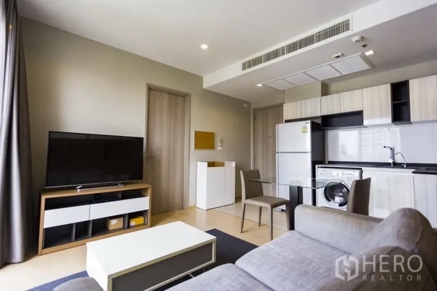 Condominium for rent in Watthana, Bangkok - Open‑plan living room with grey sofa, TV console and kitchenette with fridge and washer.