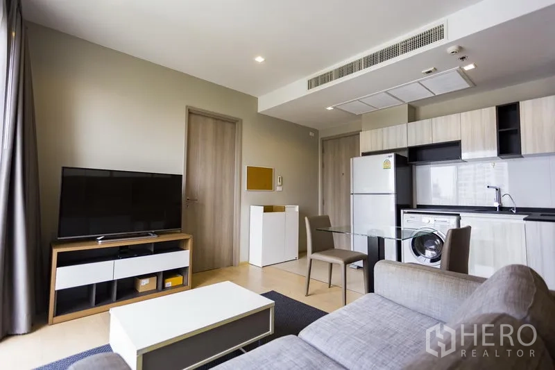 Condominium for rent in Watthana, Bangkok - Open‑plan living room with grey sofa, TV console and kitchenette with fridge and washer.
