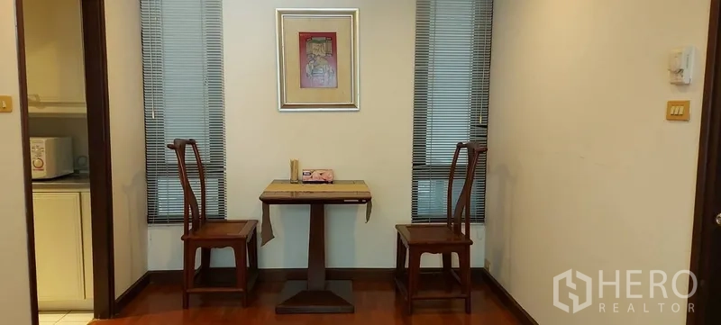 Condominium for rent in Sathon, Bangkok - Compact dining nook with two wooden chairs and a small table between narrow windows, next to the kitchenette entrance.