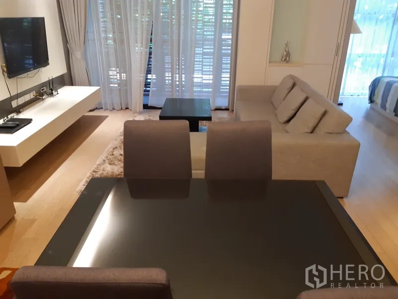公寓 出租在Watthana, Bangkok - 四人餐桌面向客厅，墙挂电视，轻薄窗帘后是绿意与阳台景致。