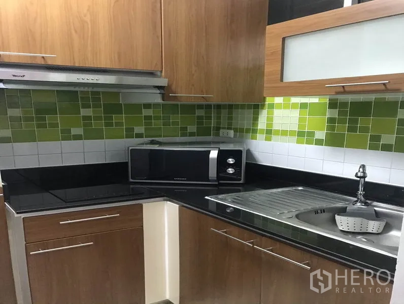 Condominium for rent or sale in Khlong Toei, Bangkok - Compact L-shaped kitchen with green mosaic backsplash, wood cabinets, black countertops, microwave and sink.