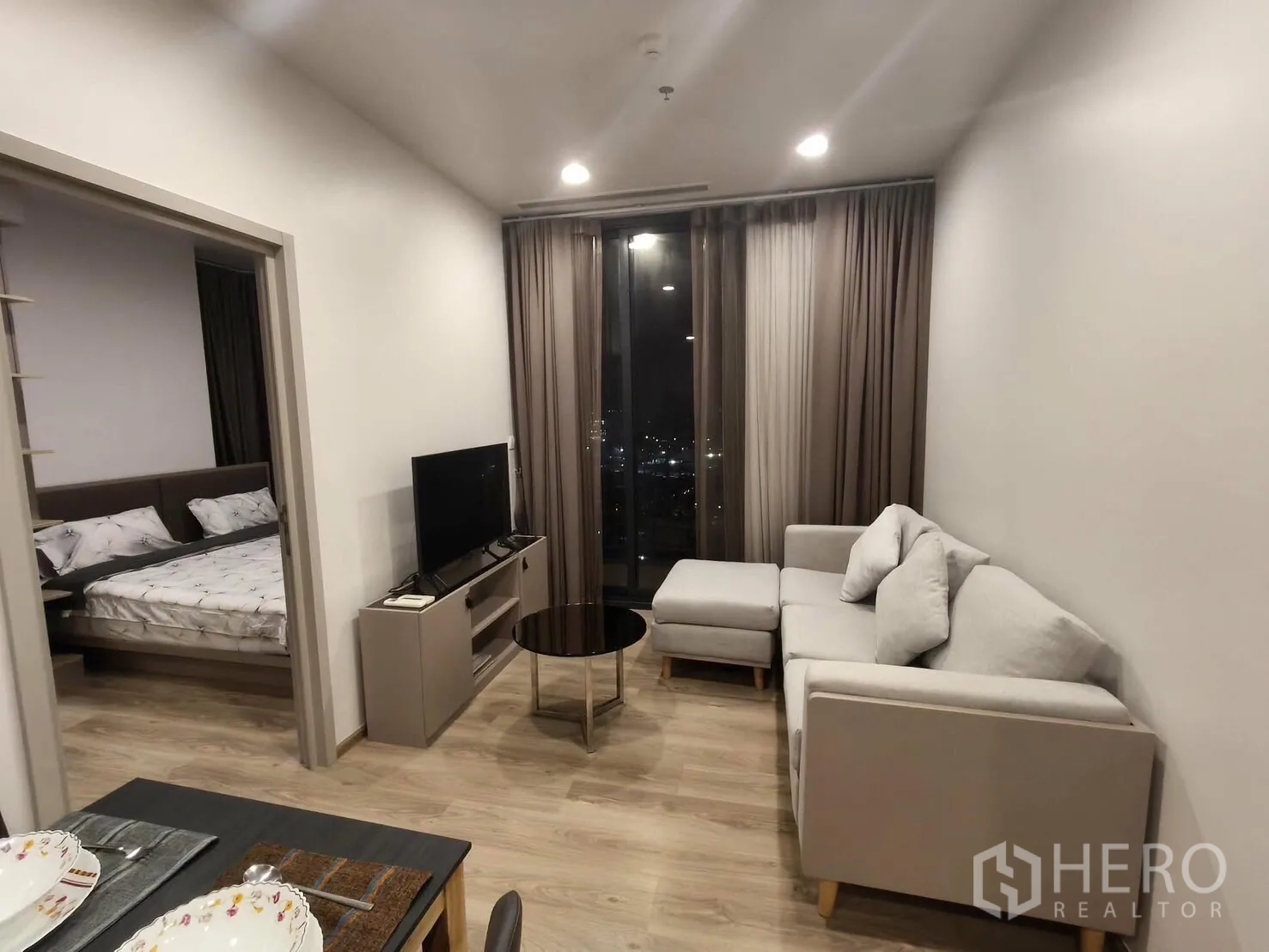 Condominium for rent in Khlong Toei, Bangkok - Cozy living room with sectional sofa, TV console, and tall curtains framing a city view, with a doorway to the bedroom.