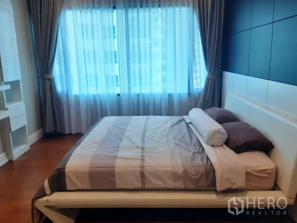 Condominium for rent in Khlong Toei, Bangkok - Bright bedroom with large window, curtains, padded headboard and wood floors in a Khlong Toei condominium.