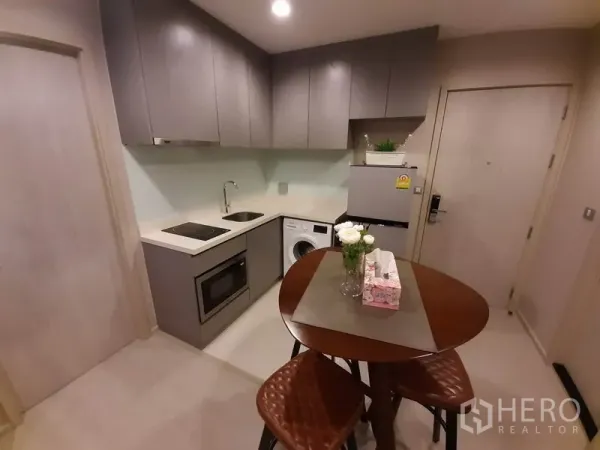 Condominium for rent or sale in Khlong Toei, Bangkok - Compact L-shaped kitchen with grey cabinets, cooktop, microwave, washer and fridge.