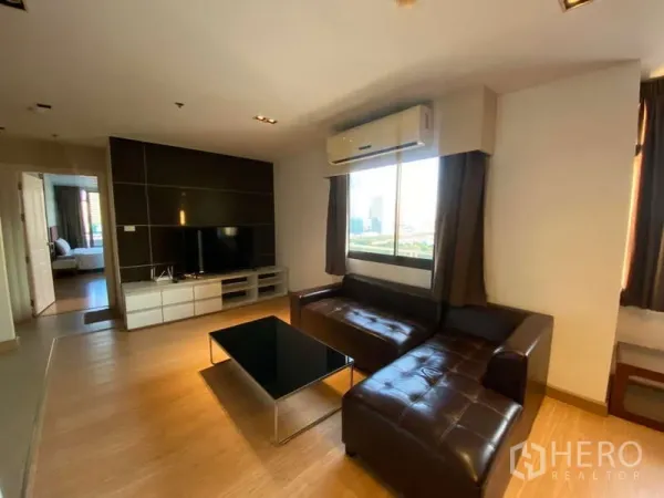 Condominium for rent in Khlong Toei, Bangkok - Bright living room with L-shaped leather sofa, TV wall unit, wood flooring and window with city view.