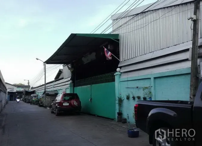 Warehouse for rent in Bang Kho Laem, Bangkok - Metal-clad warehouse row with green perimeter wall, covered loading bay, Thai flag, and narrow access road with parked vehicles in Bang Kho Laem.