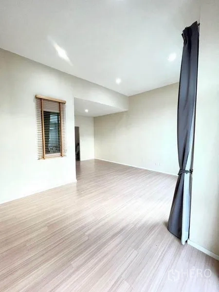 Townhouse for sale in Bang Kapi, Bangkok - Large empty room with light wood floors, a window with blinds, and tall curtains.