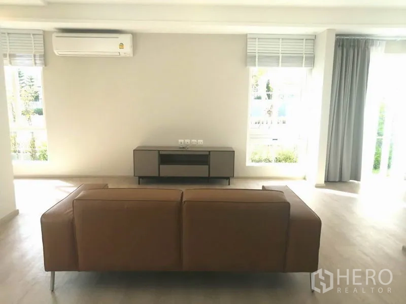 Townhouse for rent in Prawet, Bangkok - Bright living room with large windows, air conditioner, light wood floors and a leather sofa facing a low TV console.